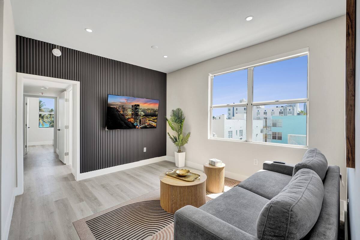 A modern living room with a grey sofa, a flat-screen TV mounted on a dark wall, a potted plant, and a view of buildings through a large window.