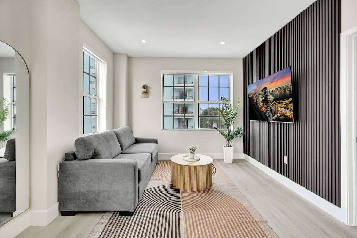 A modern living room with a grey sofa, a round wooden coffee table, a large flat-screen TV mounted on a dark wall, and a large window with a city view.