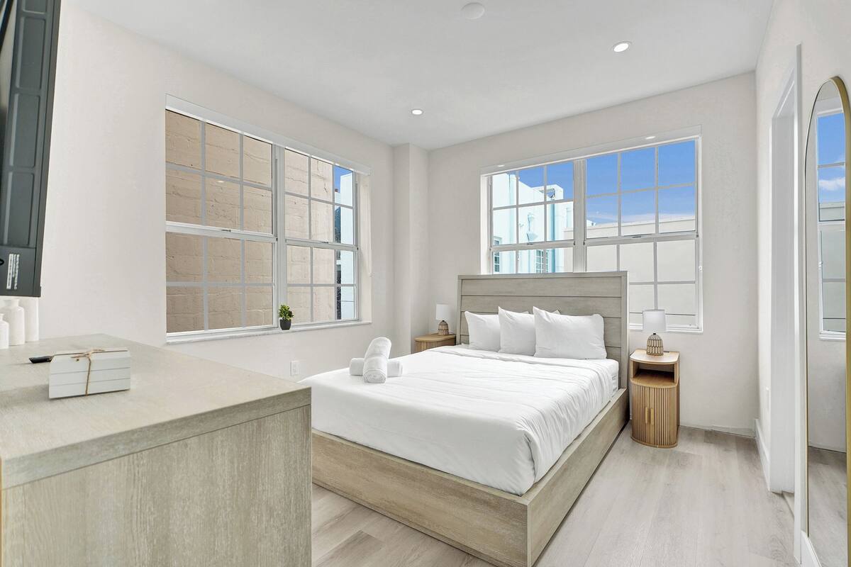 A modern hotel room with a large bed, two bedside tables, a small desk, and large windows.