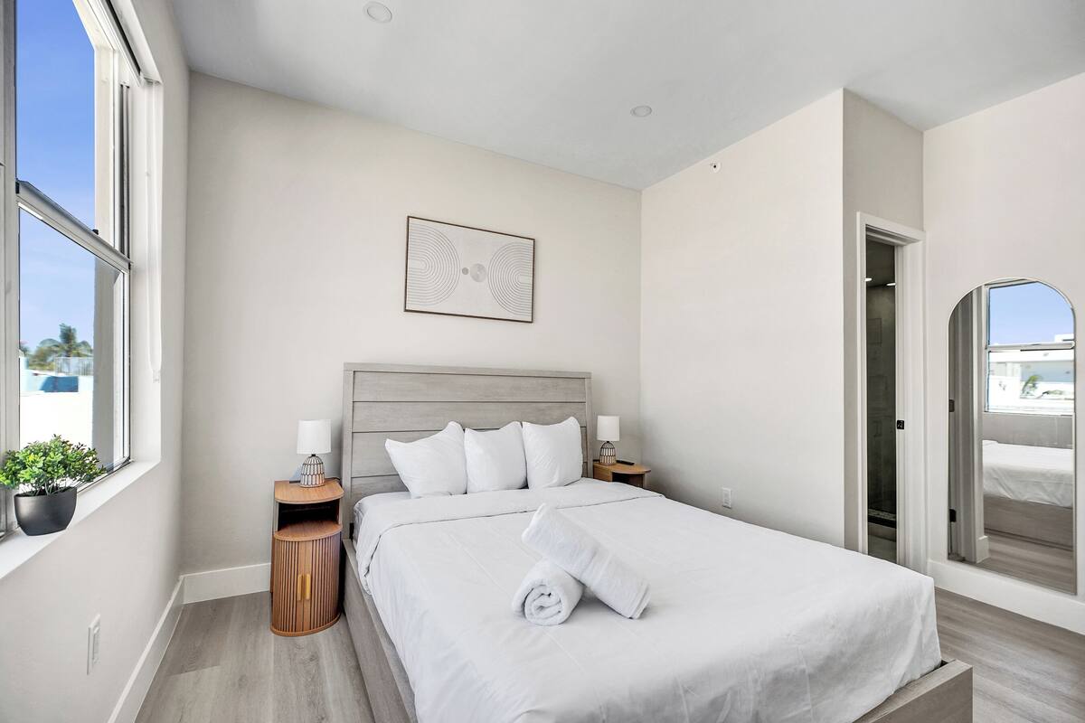 A modern hotel room with a large bed, a bedside table, a nightstand, a potted plant, and a view through an open door.