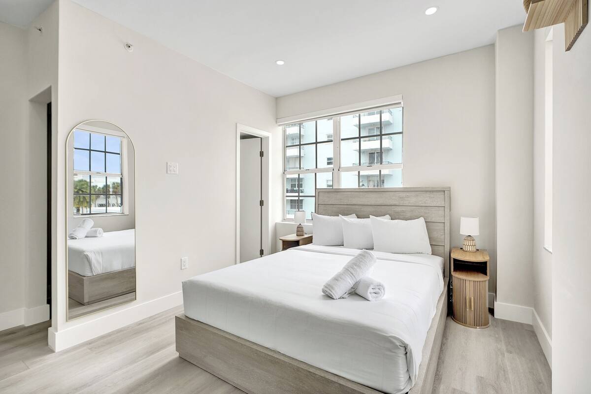 A modern hotel room with a large bed, a mirror, a nightstand, and a window with a view of buildings.
