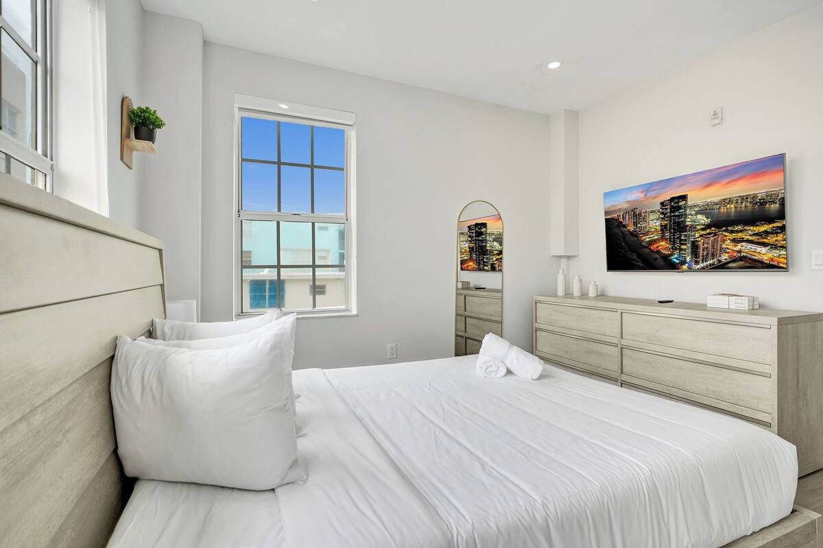 A bedroom with a bed, a dresser, a window, and a cityscape painting.