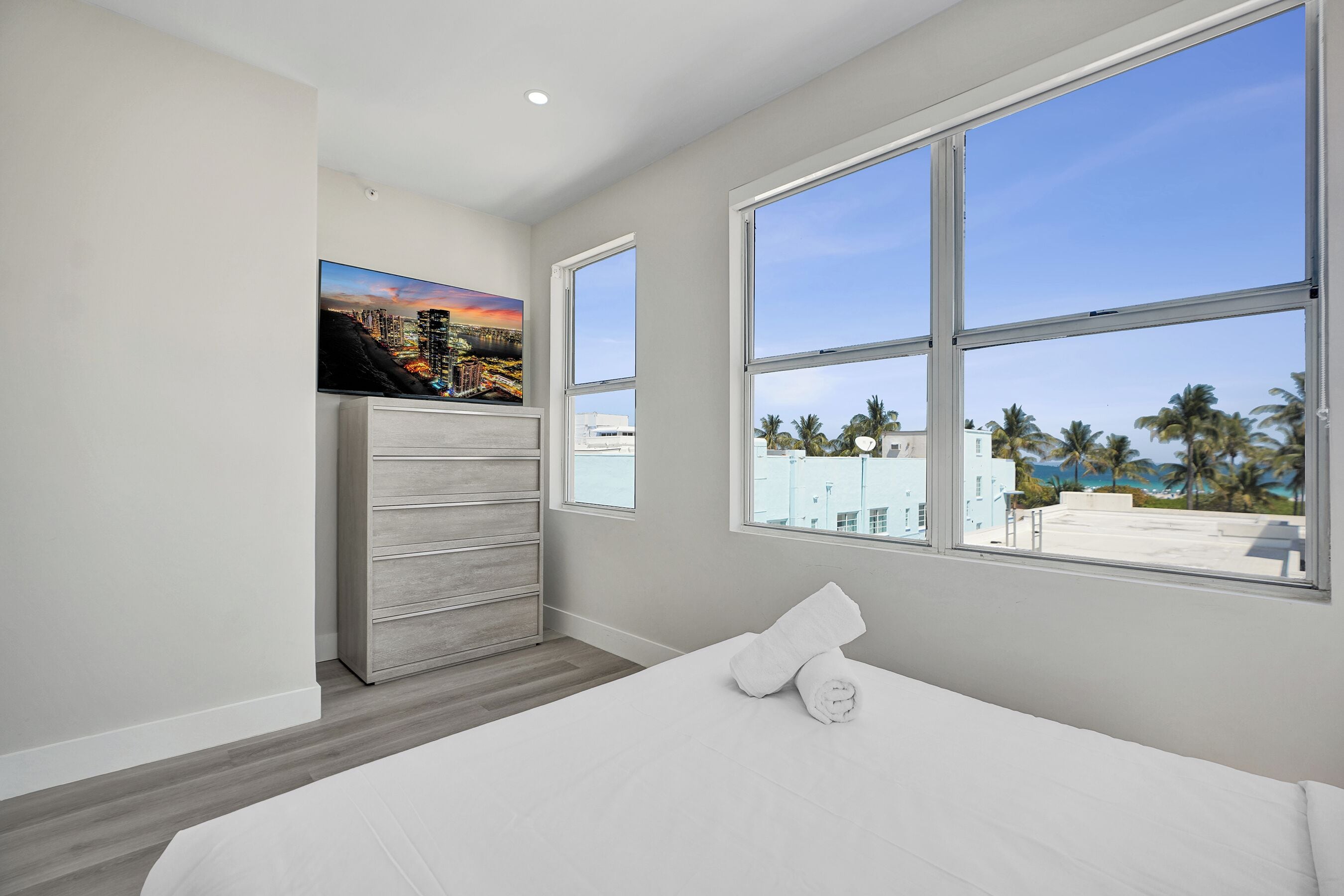 A bedroom with a bed, a dresser, a painting, and a view of palm trees and buildings through large windows.