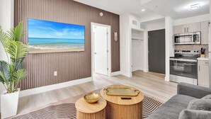 A modern living room with a large flat-screen TV, a wooden wall panel, a kitchen area with a microwave, and a dining area with a bowl and tray. - SoBe Suites 727 (Miami Beach)