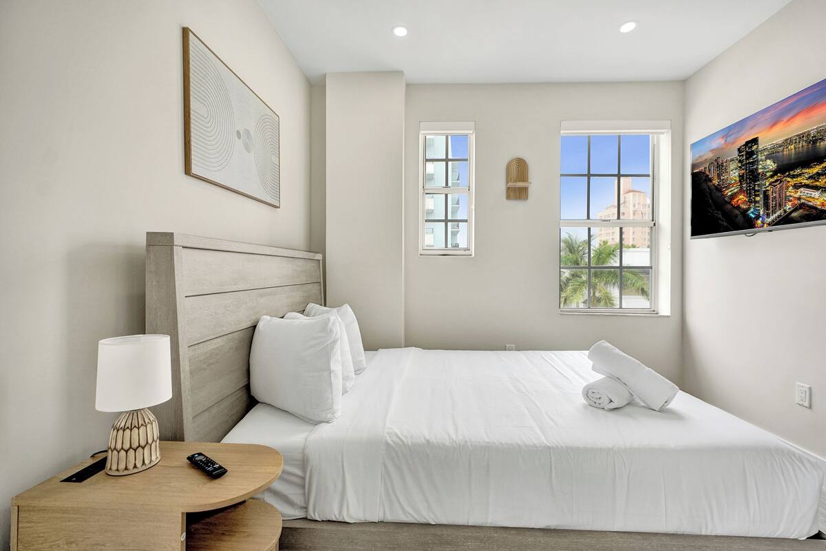 A hotel room with a bed, a nightstand with a lamp, a remote control, a window with a view of palm trees, and a painting on the wall.