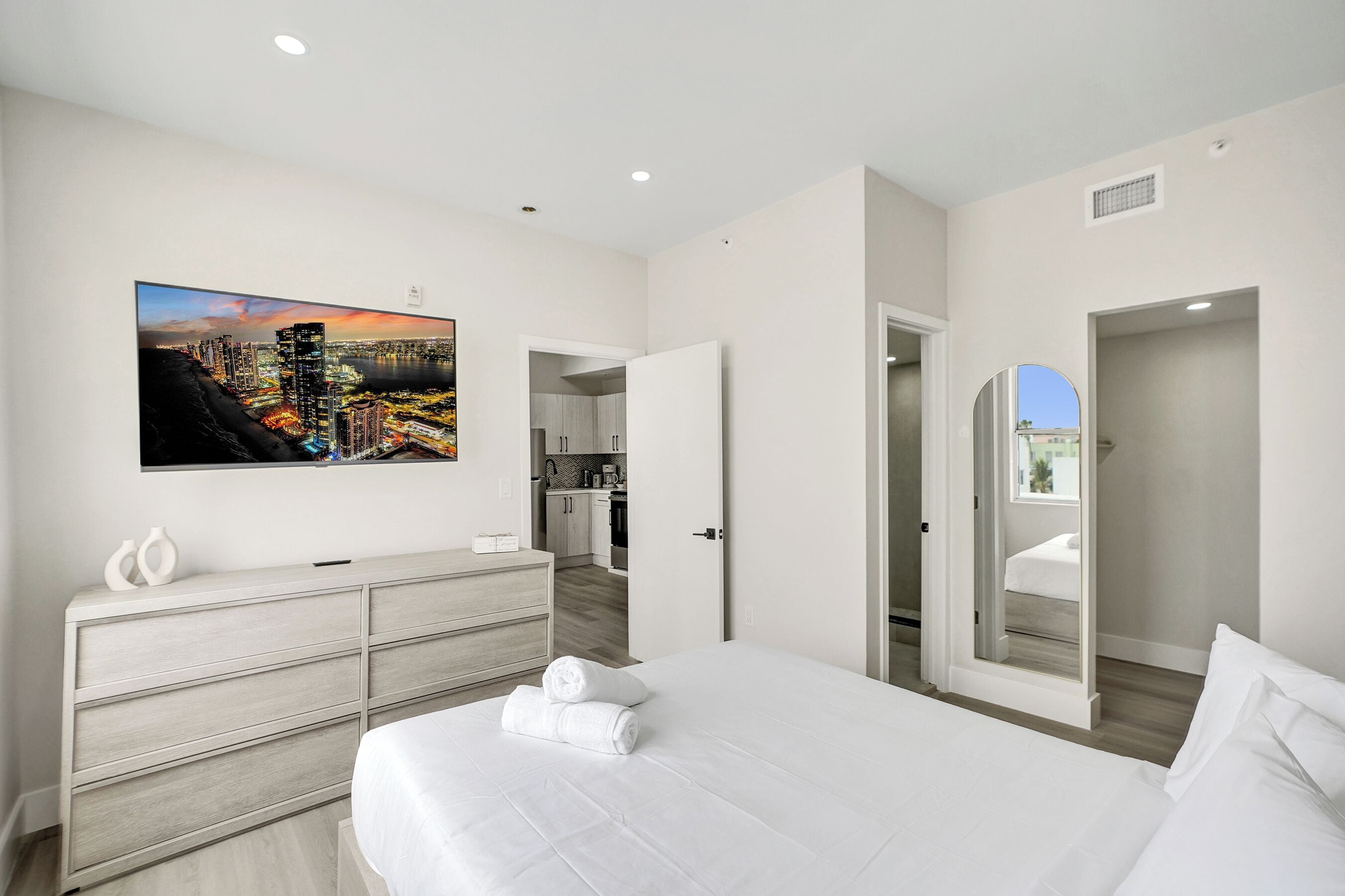 Habitación de hotel moderna con una cama grande, un aparador, un cuadro con una vista urbana, y vistas a la cocina y al balcón.