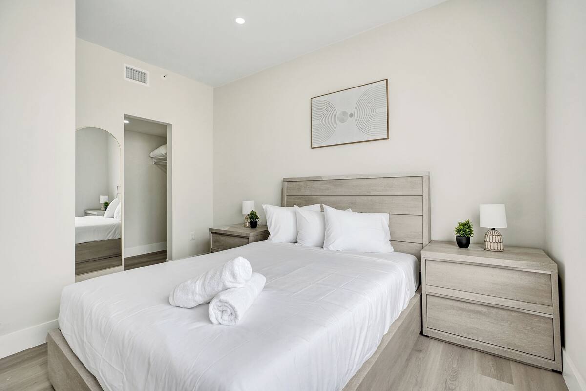 A modern hotel room with a large bed, a nightstand, a mirror, and a framed artwork on the wall.