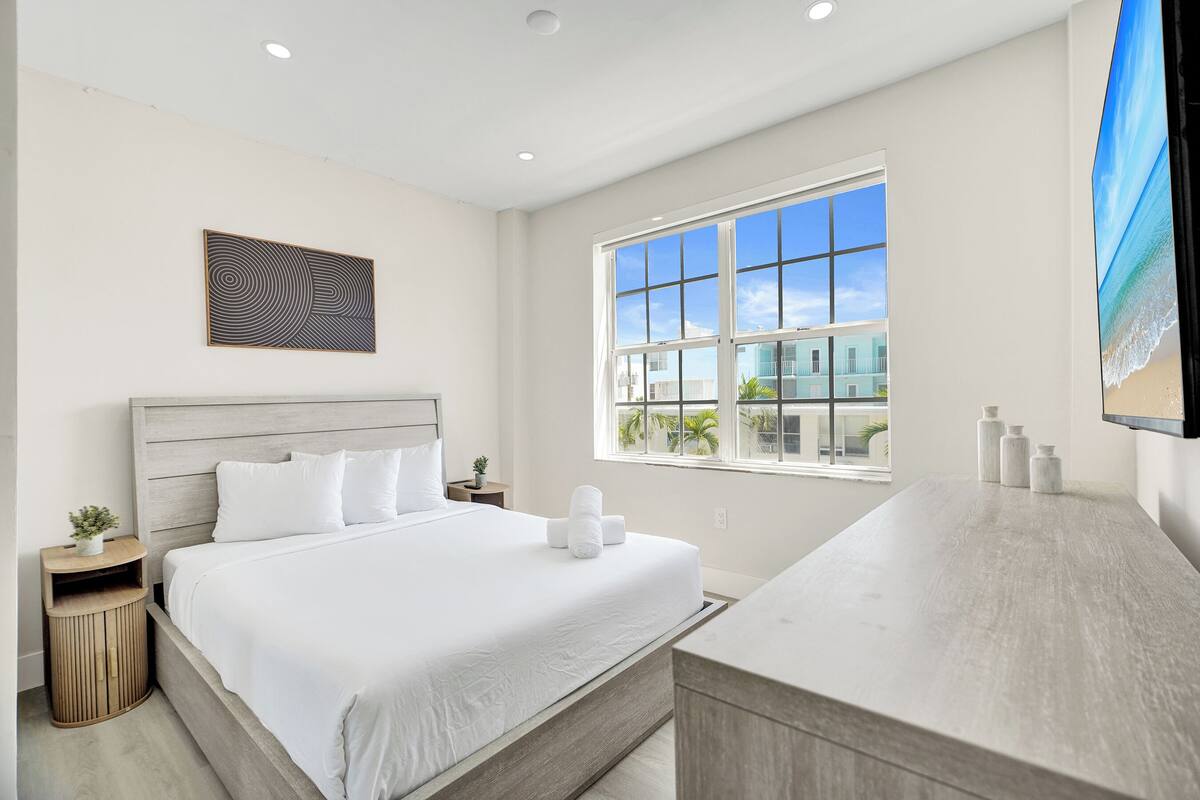 A modern bedroom with a large bed, a wooden headboard, a window with a view of buildings, and a mounted TV.