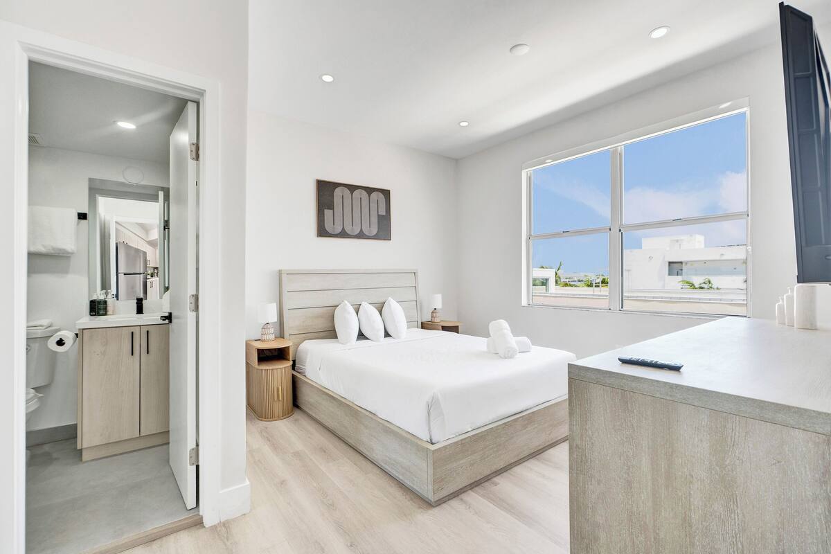 A modern hotel room with a large bed, a bedside table, a bathroom with a toilet and sink, and a view of buildings through a large window.