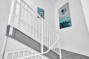 Un escalier blanc avec une rampe, un mur orné de trois tableaux encadrés et une porte en bas.