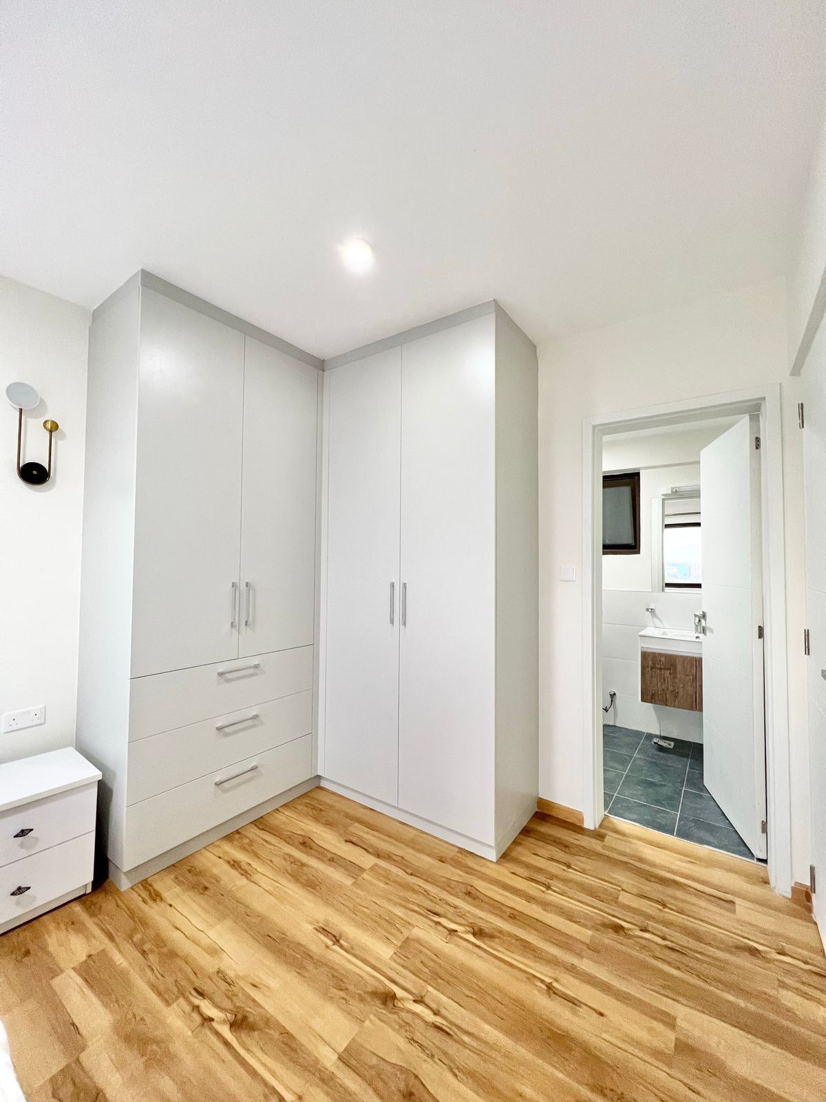 Een kamer met een grote witte garderobe, een klein wit nachtkastje en een houten vloer.