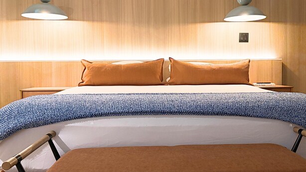 Una cama bien hecha con una colcha azul y blanca estampada, dos almohadas marrones y dos lámparas colgantes encima.
