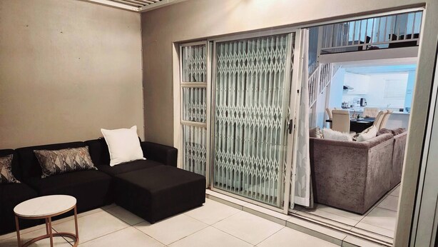 A living room with a black sofa, a white pillow, a small round table, and a sliding glass door leading to a balcony with a chair. - Luxury Apartment in Waikiki E5 Close to the Beach (Ballito)