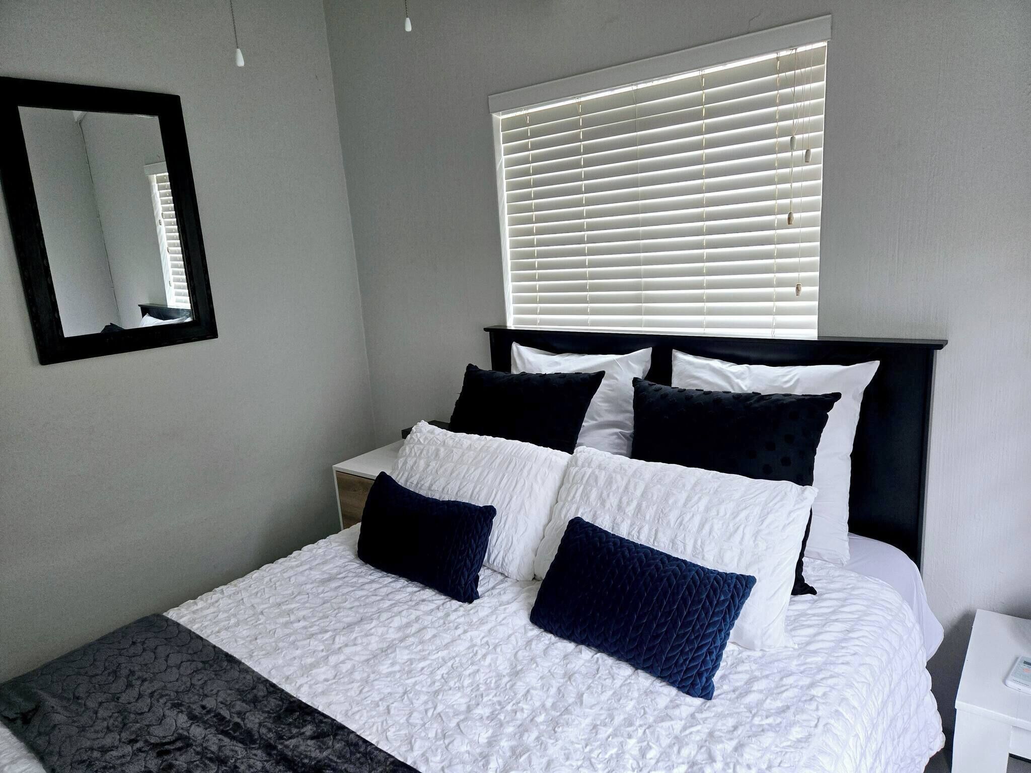 A bedroom with a bed, a bedside table, a nightstand, a window with blinds, and a mirror on the wall.