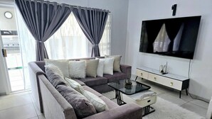 A modern living room with a sofa, a glass coffee table, a white furry rug, a TV, and a white cabinet. - Luxury Apartment in Waikiki E5 Close to the Beach (Ballito)