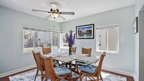A dining area with a glass table, wicker chairs, a ceiling fan, and large windows. - 3bed 2bath beach cottage Steps to the Sand in MB (San Diego)