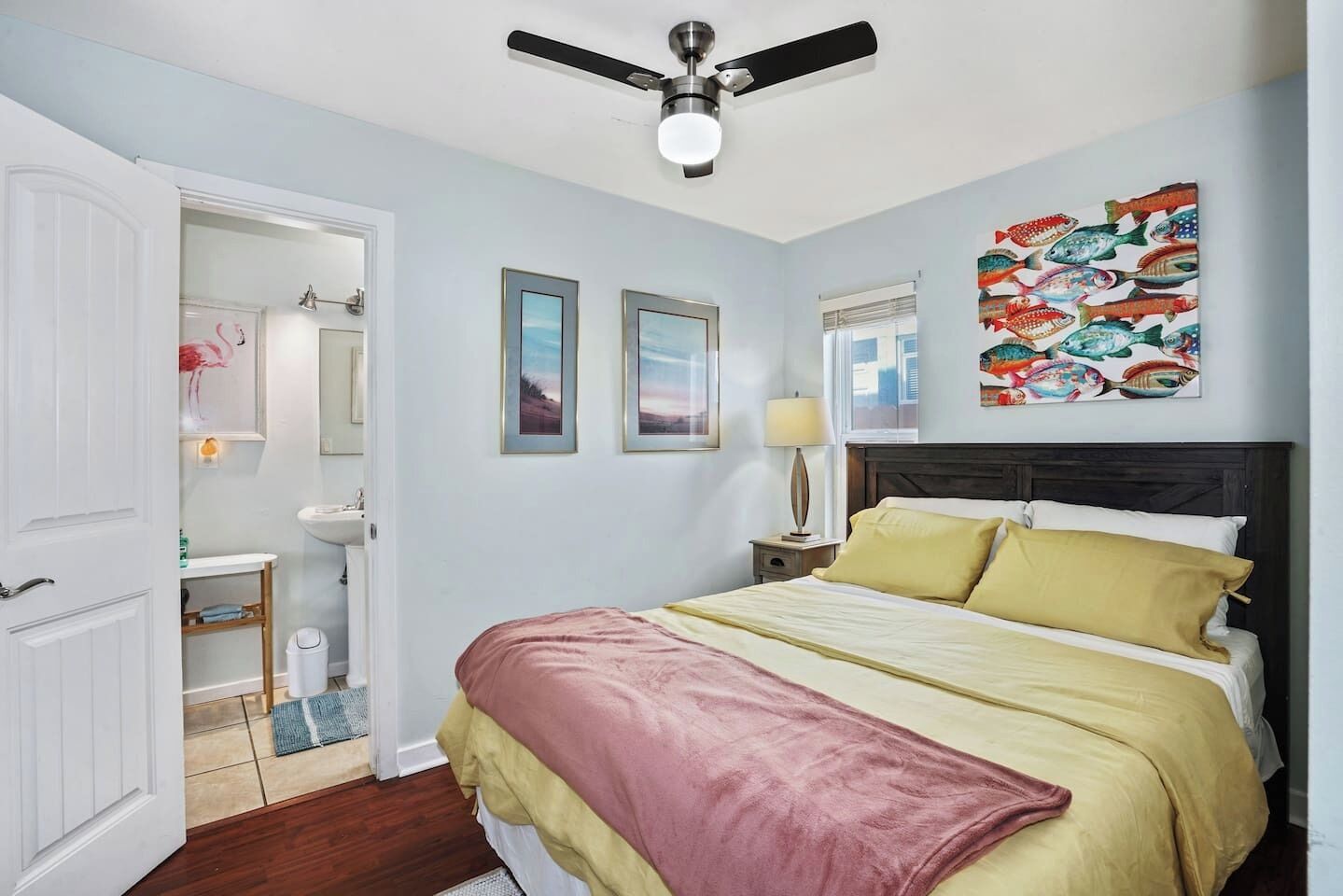 A bedroom with a bed, a ceiling fan, a bathroom with a sink, and a wall with framed pictures.