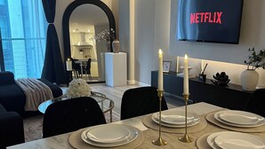 A modern living room with a dining area set for six, a flat-screen TV displaying 'NETFLIX,' and a view of the kitchen and balcony. - Liverpool City Centre Luxury 2 Bedroom 2 Bathroom Balcony Flat - Cheap Parking (Liverpool)