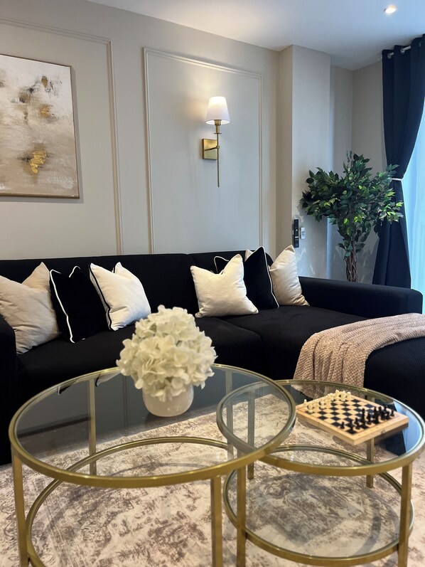 A modern living room with a black sofa, white cushions, a glass coffee table, a chessboard, and a potted plant.
