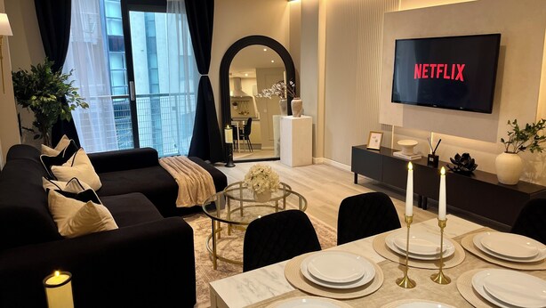 A modern living room with a sofa, dining table set for four, a TV displaying 'NETFLIX,' and a view into the kitchen. - Liverpool City Centre Luxury 2 Bedroom 2 Bathroom Balcony Flat - Cheap Parking (Liverpool)