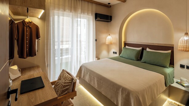 Una habitación de hotel con cama, escritorio con computadora portátil, silla, perchero y aire acondicionado de pared.