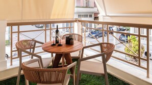 Un balcón con una mesa redonda y cuatro sillas de mimbre, barandilla blanca y vistas a coches estacionados y edificios.