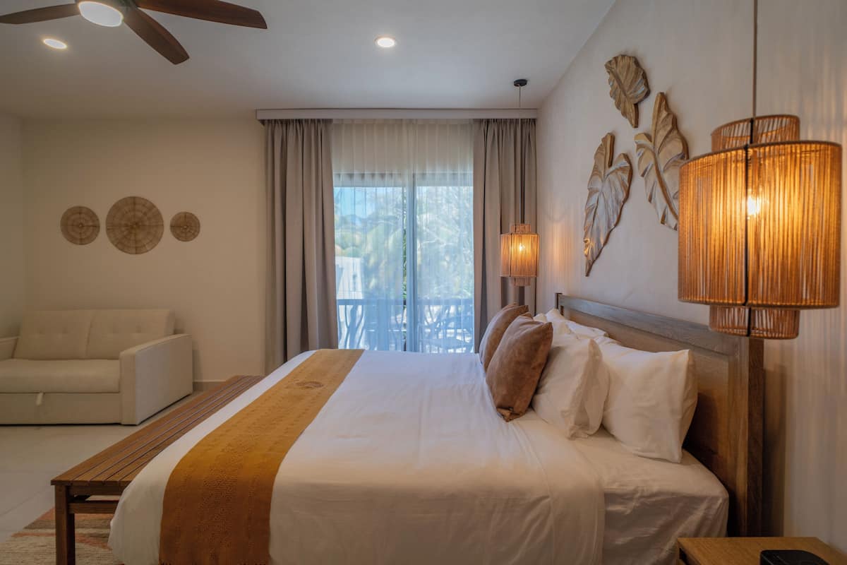 A hotel room with a bed, a ceiling fan, a nightstand, a lamp, and a view of the outdoors.