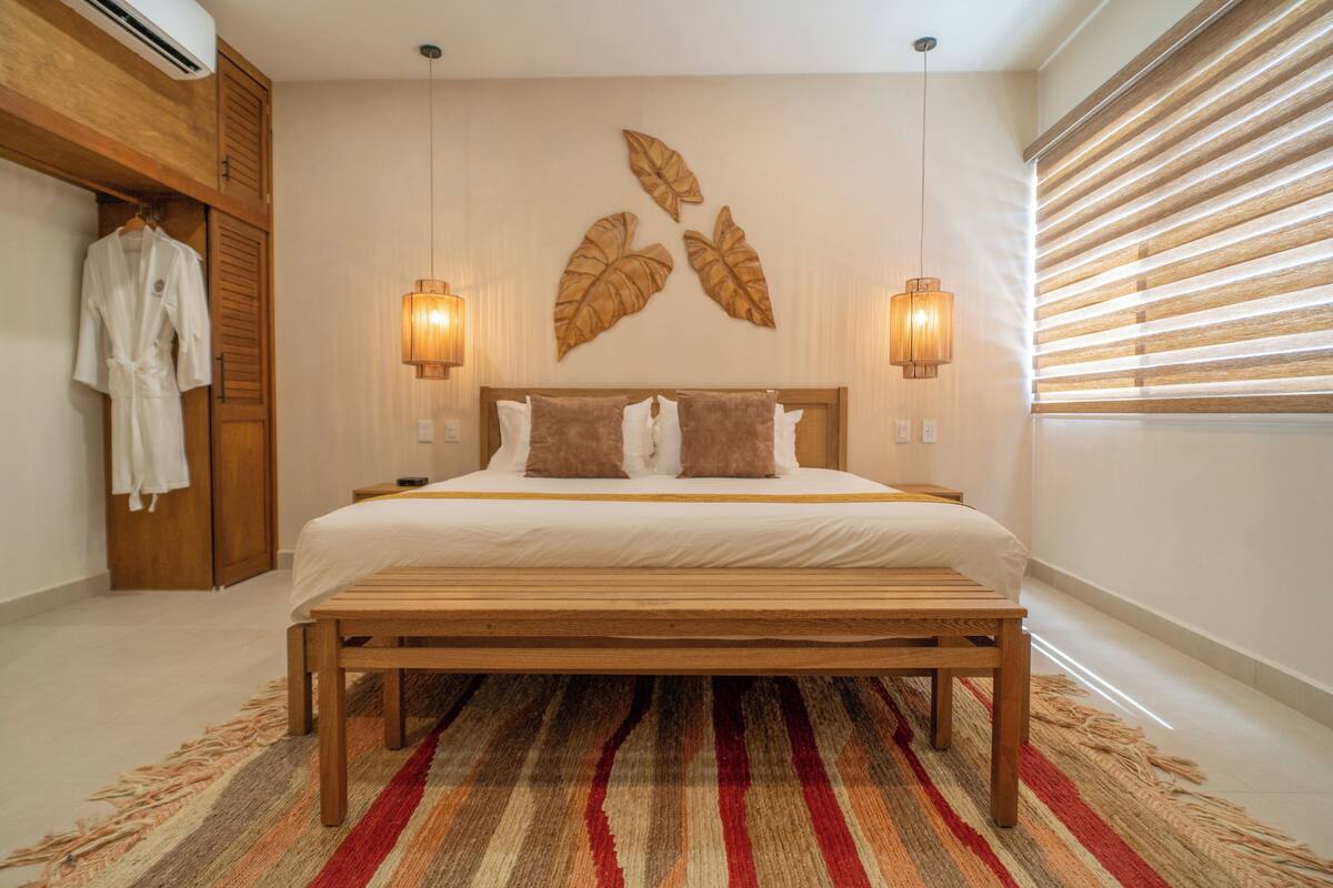 A bedroom with a wooden bed, a bench, a large wall art piece, and two wall-mounted lamps.