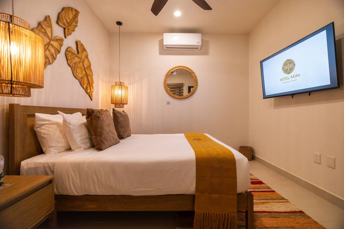 A hotel room with a bed, a wall-mounted TV displaying the hotel name, a ceiling fan, and a round mirror.