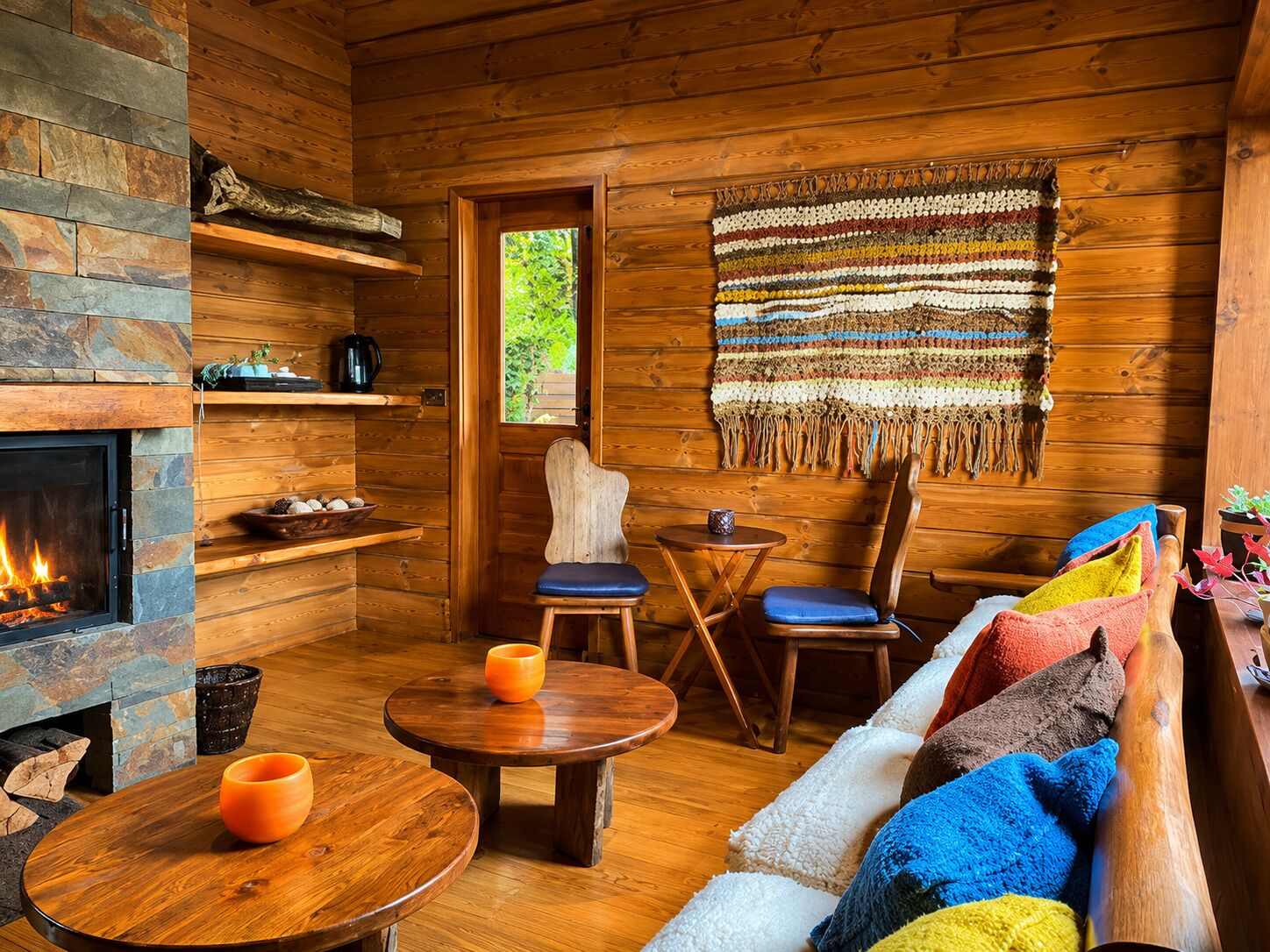 A cozy cabin interior with a fireplace, wooden walls, a woven rug, and a sofa with colorful cushions.