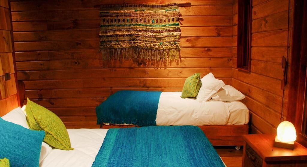 Une chambre dans un chalet en bois, avec deux lits, une couverture tissée suspendue au mur et une table de chevet avec une lampe.