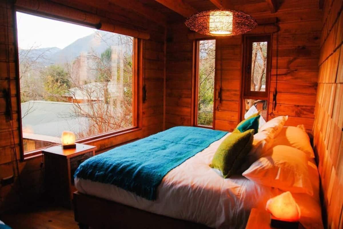 Un dormitorio de cabaña de madera con una cama, una mesita de noche, una lámpara y una ventana con vistas a montañas.