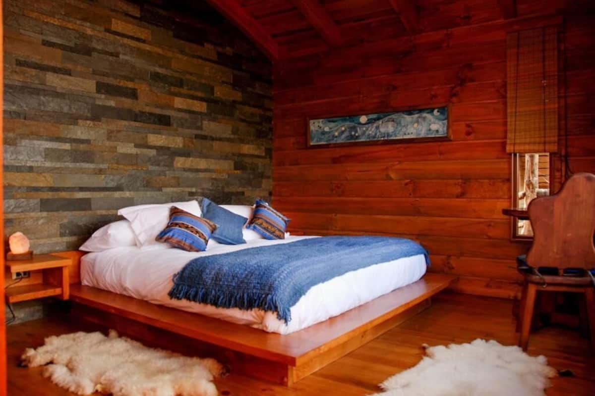 Un dormitorio de cabaña de madera con una cama, almohadas, un edredón, un cuadro y una silla.