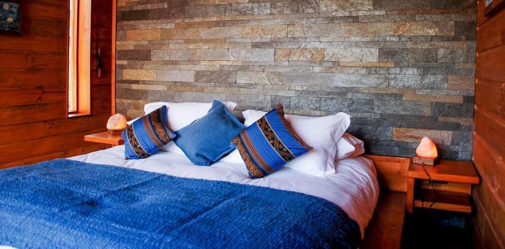 A wooden cabin-style bedroom with a stone wall, a bed with blue and patterned pillows, and two bedside lamps.