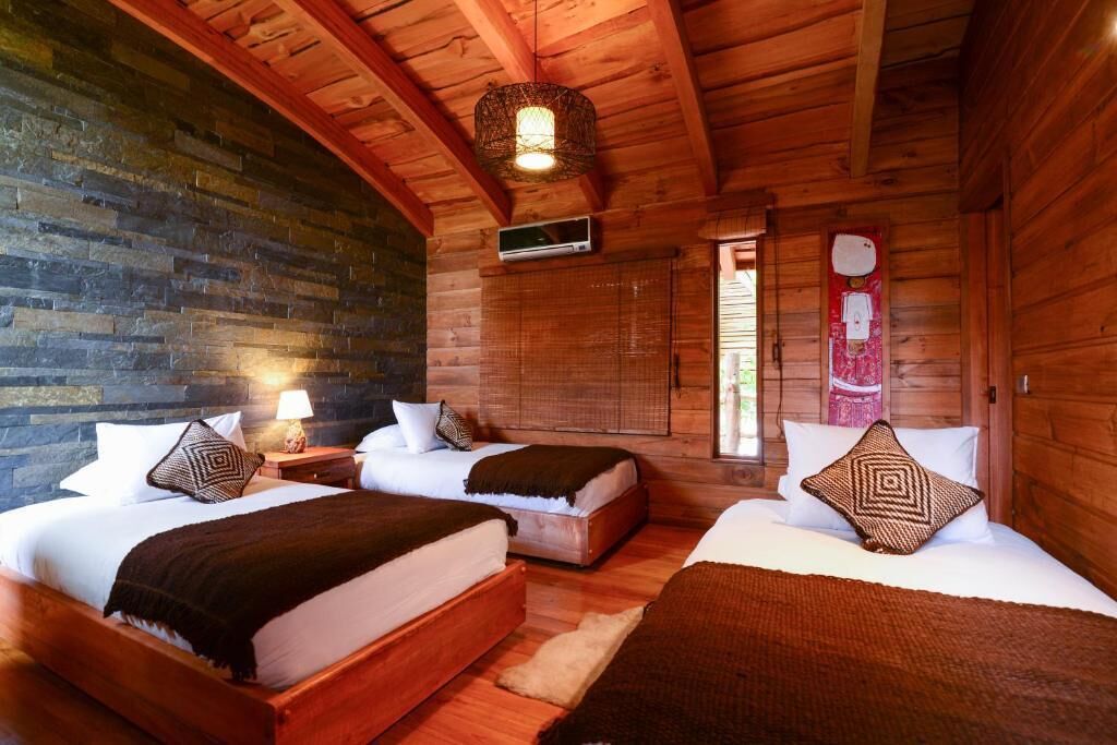 A log cabin room with two beds, a stone wall, wooden ceiling beams, and a hanging light fixture.