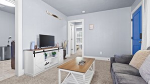Sala de estar com televisão, móvel de armazenamento branco, mesa de centro de madeira, sofá cinza e porta azul.