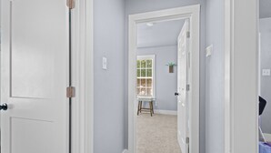 Um corredor com portas brancas, uma janela com persianas, um banquinho pequeno e uma planta na parede.