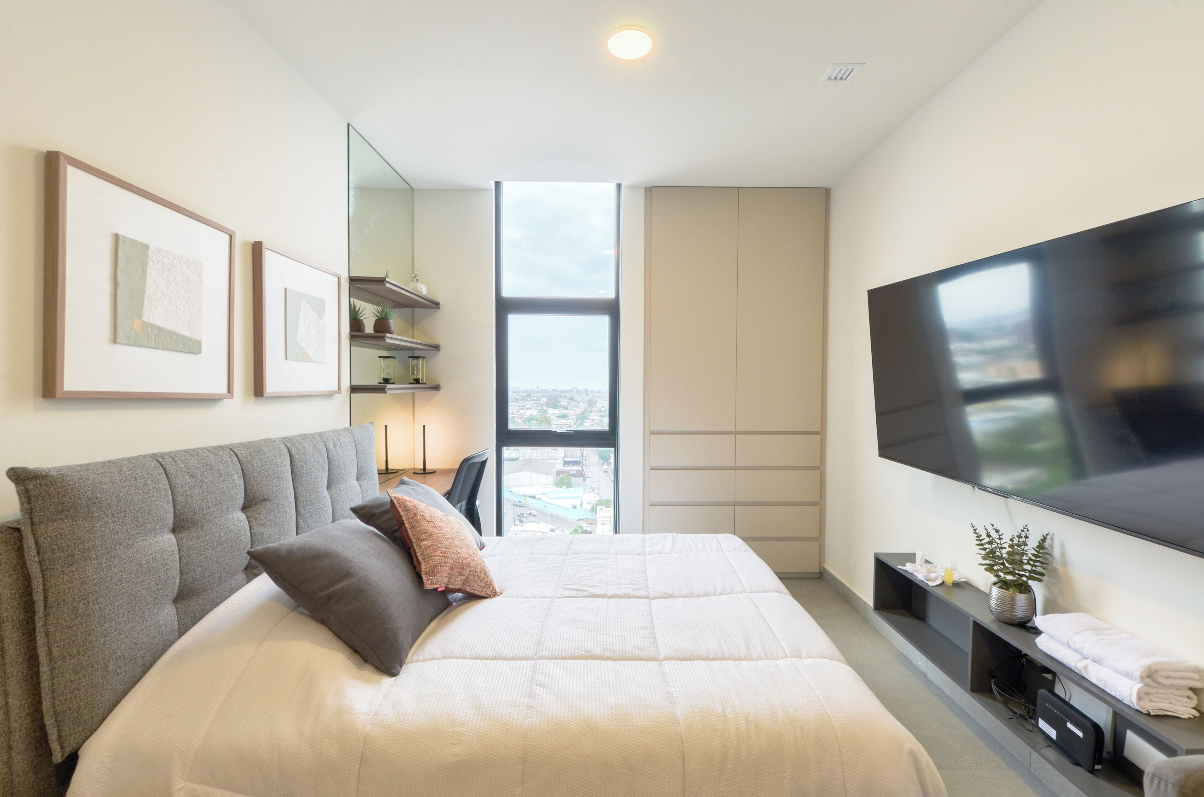 A modern bedroom with a large bed, a flat-screen TV, a window with a city view, and framed artwork on the wall.