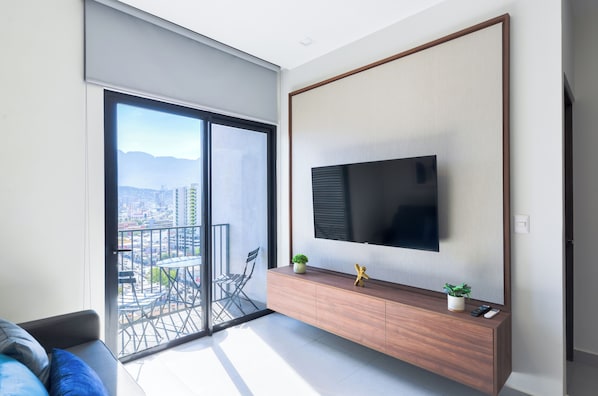 A modern living room with a large TV, a sliding glass door to a balcony, and a minimalist wooden console. - Colonia (Monterrey)