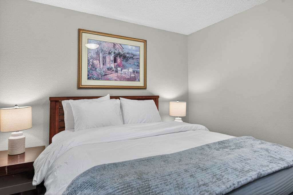 A hotel room with a bed, two bedside lamps, a framed painting on the wall, and a window.