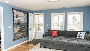 Ein Wohnzimmer mit einem grauen Ecksofa, einem großen Wandteppich und Blick durch die Tür.