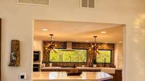 A modern kitchen with a large island, two pendant lights, and a view of greenery through the windows. - Ultimate AFA Graduation Estate: 5 Private Acres - 5 Mins to North Gate (Colorado Springs)