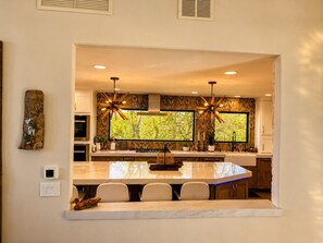 Una cocina moderna con una isla grande, dos lámparas colgantes y vistas a la vegetación por las ventanas.
