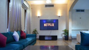 Ein Wohnzimmer mit großem Flachbildfernseher an der Wand, einem blaugrünen Sofa, einem Couchtisch und zwei Blumentöpfen.