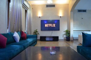 Ein Wohnzimmer mit großem Flachbildfernseher an der Wand, einem blaugrünen Sofa, einem Couchtisch und zwei Blumentöpfen.