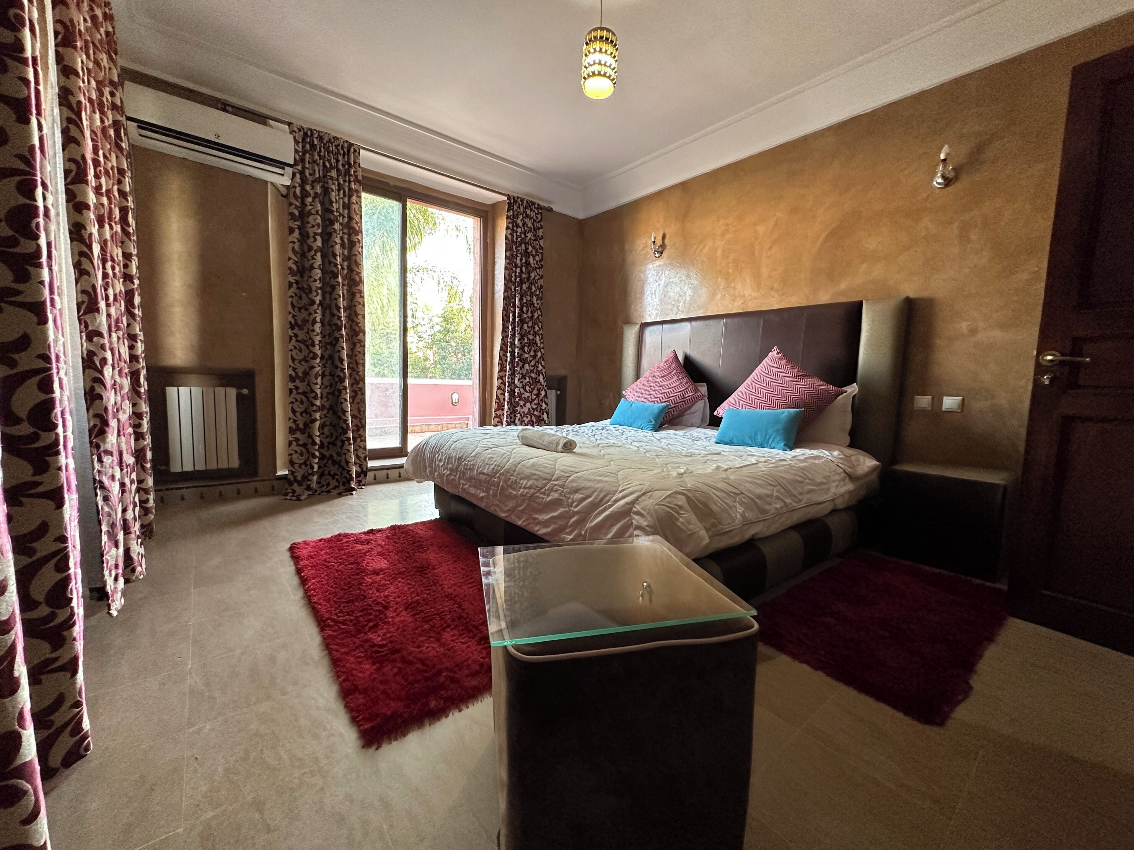 Une chambre à coucher avec un lit, deux tables de chevet, un tapis rouge, une table avec un plateau en verre et une fenêtre avec des rideaux.