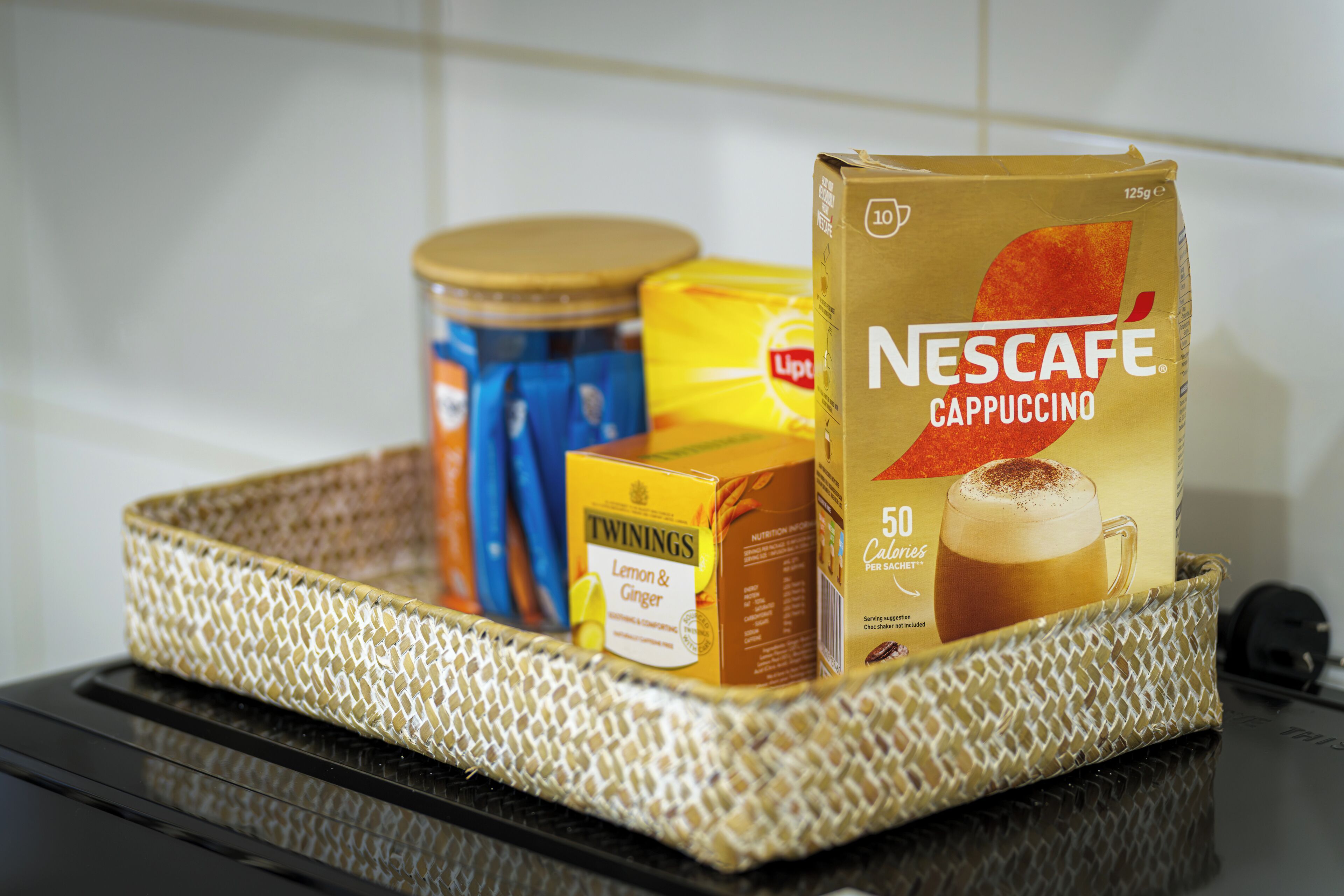 Una encimera de cocina con una cafetera, una lata de café, una caja de té Twinings y un sobre de cappuccino Nescafé.