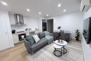 Eine moderne Küche mit weißen Schränken, einem grauen Sofa, einem Essbereich mit Tisch und einem an der Wand befestigten Fernseher.