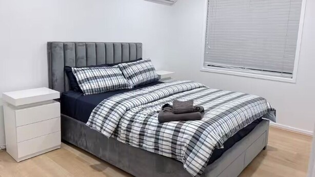 Un dormitorio con una cama tapizada en gris, mesitas de noche blancas, una colcha a cuadros y una ventana con persianas.