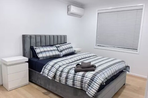 Un dormitorio con una cama tapizada en gris, mesitas de noche blancas, una colcha a cuadros y una ventana con persianas.